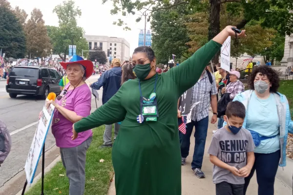 Photo of person of color wearing a green dress and face mask, with arms spread in joy