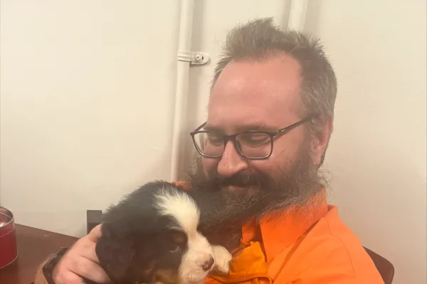Photo of white man with short dark hair and beard in an orange shirt, holding a puppy