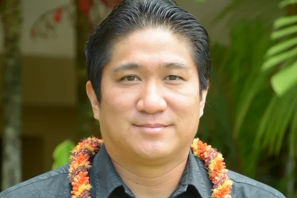 Photo of man with short dark hair and dark button down shirt and wearing an orange lei