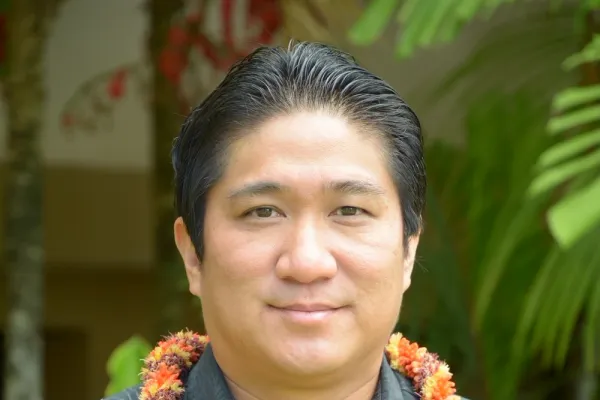 Photo of man with short dark hair and dark button down shirt and wearing an orange lei
