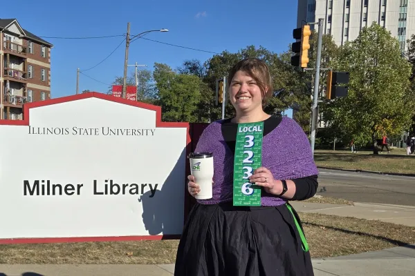 Photo of woman in a floor length black dress and purple knit cabled shawl in front of Milner Library sign
