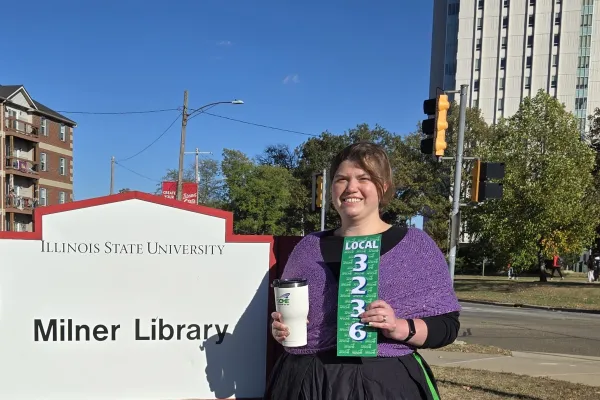 Photo of woman in a floor length black dress and purple knit cabled shawl in front of Milner Library sign