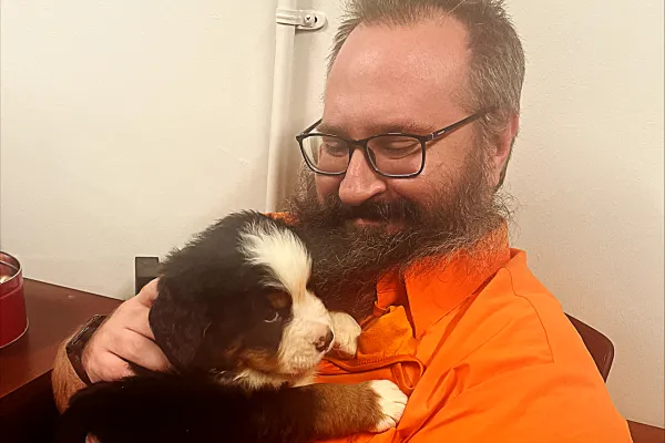 Photo of white man with short dark hair and beard in an orange shirt, holding a puppy