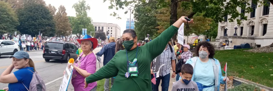 Photo of person of color wearing a green dress and face mask, with arms spread in joy