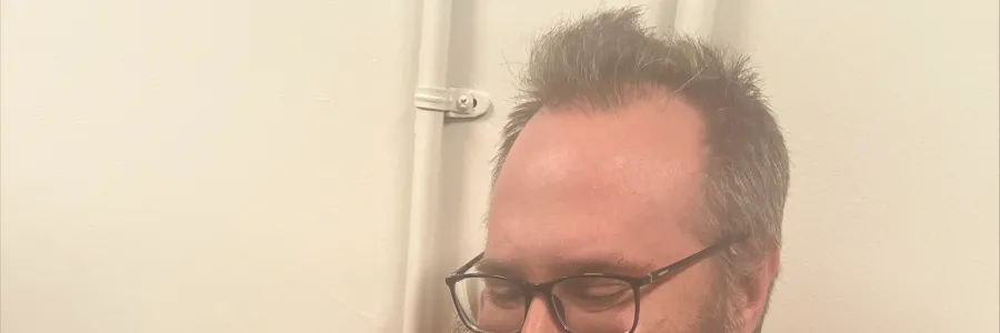 Photo of white man with short dark hair and beard in an orange shirt, holding a puppy