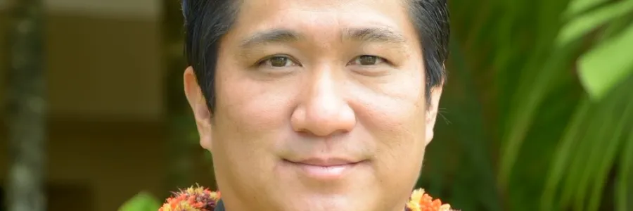 Photo of man with short dark hair and dark button down shirt and wearing an orange lei
