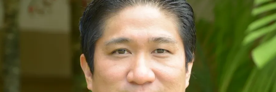 Photo of man with short dark hair and dark button down shirt and wearing an orange lei