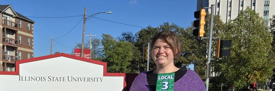 Photo of woman in a floor length black dress and purple knit cabled shawl in front of Milner Library sign