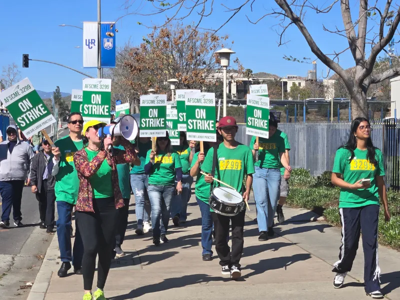 AFSCME Members in green shirts walking picket line with signs that say ON STRIKE.