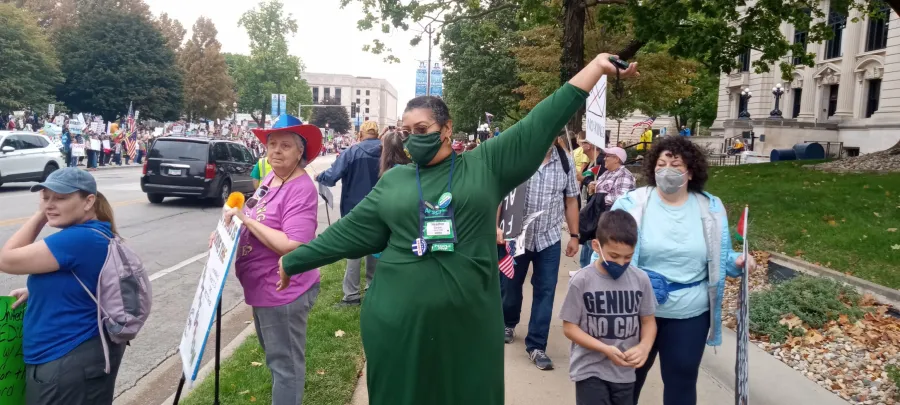 Photo of person of color wearing a green dress and face mask, with arms spread in joy