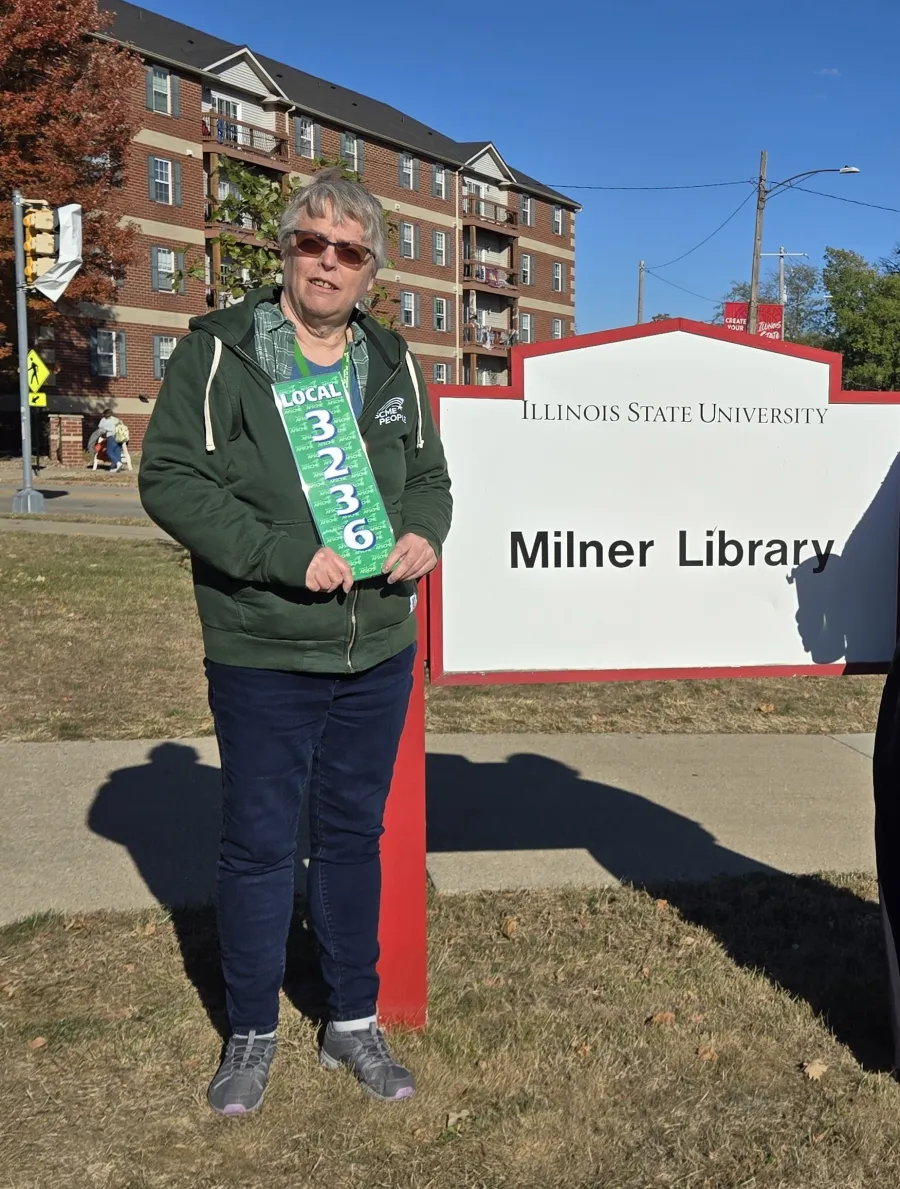 Photo of woman with short grey hair standing in front of Milner Library sign holding an AFSCME Local 3236 sign