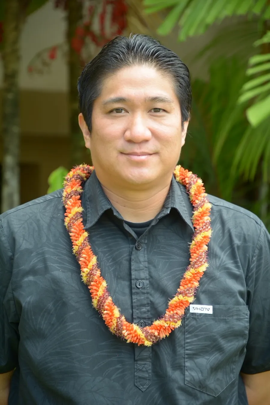 Photo of man with short dark hair and dark button down shirt and wearing an orange lei