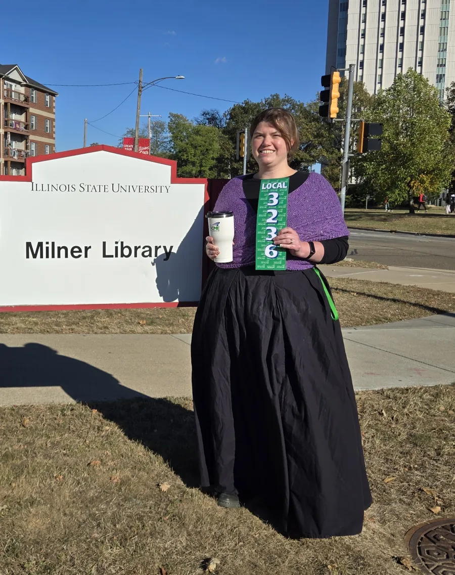 Photo of woman in a floor length black dress and purple knit cabled shawl in front of Milner Library sign
