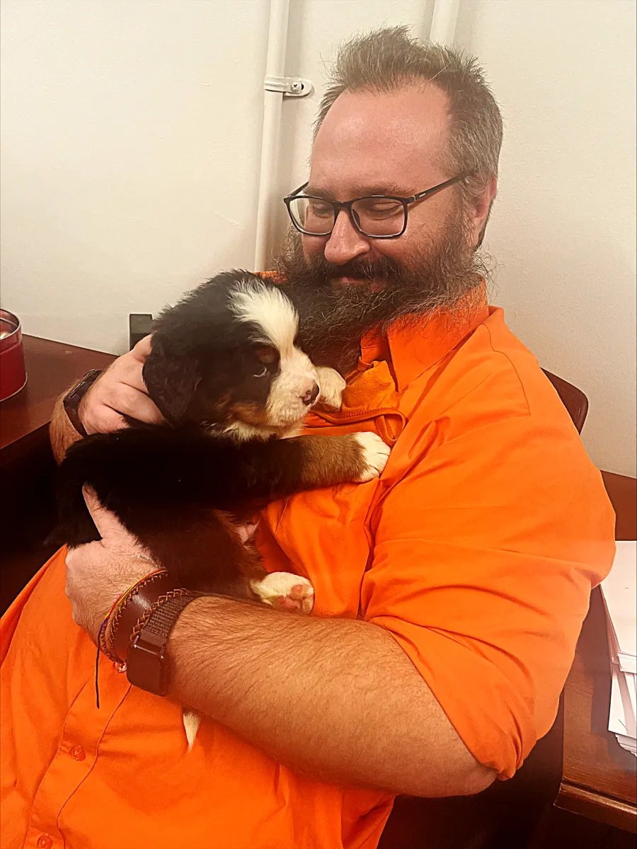 Photo of white man with short dark hair and beard in an orange shirt, holding a puppy